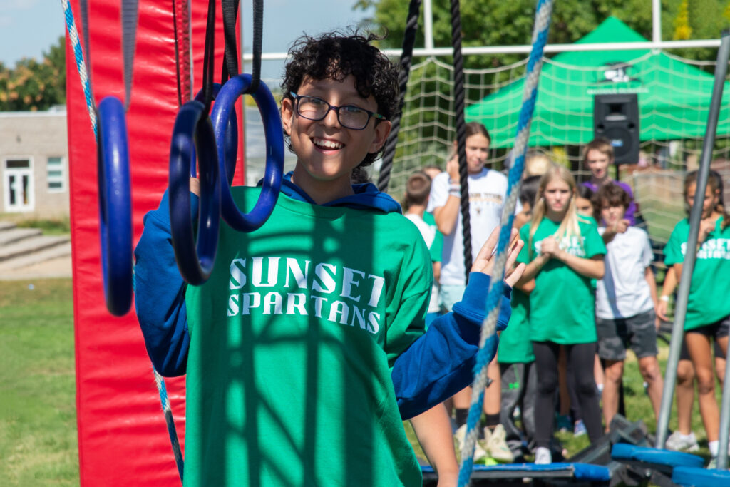 Male middle school student smiling and wearing a Sunset Spartans t-shirt and glasses. He has short curly hair and is standing in front of swinging rings waiting for his turn. A group of students is standing in the background behind him. 