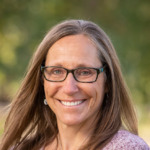 Portrait of female principal with long hair smiling for camera with blurred green trees in the background.