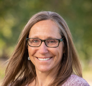 Portrait of female principal with long hair smiling for camera with blurred green trees in the background.