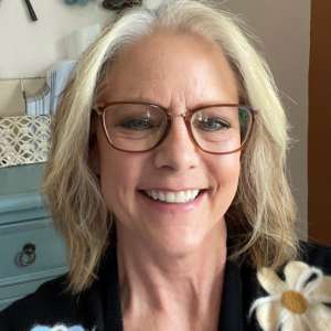 A woman with medium-length gray hair smiles while wearing glasses and a black cardigan with a flower design.