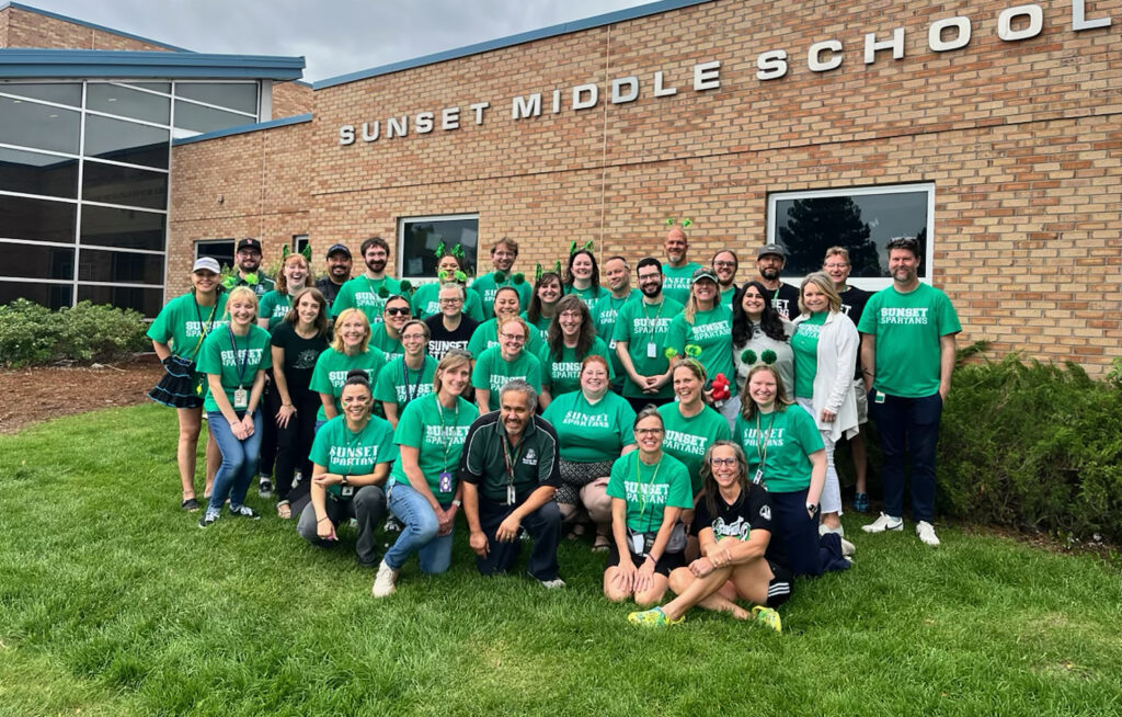 Group photo of staff members in green "Sunset Spartans" shirts outside Sunset Middle School, smiling and posing together.