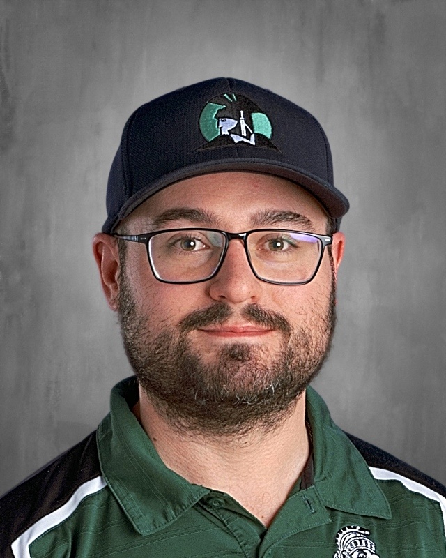 Man with glasses and a beard wearing a dark cap and green shirt, facing forward against a gray background.