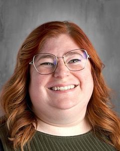 Smiling person with red hair and glasses against a gray background.