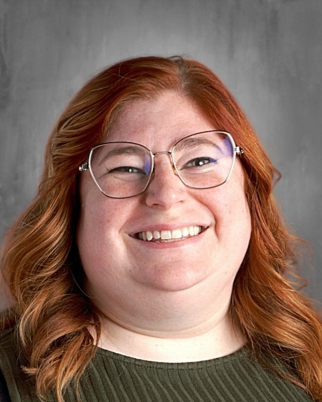 Smiling person with red hair and glasses against a gray background.