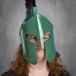 Persona con el pelo largo que lleva un casco espartano verde con un penacho negro, sobre un fondo gris.