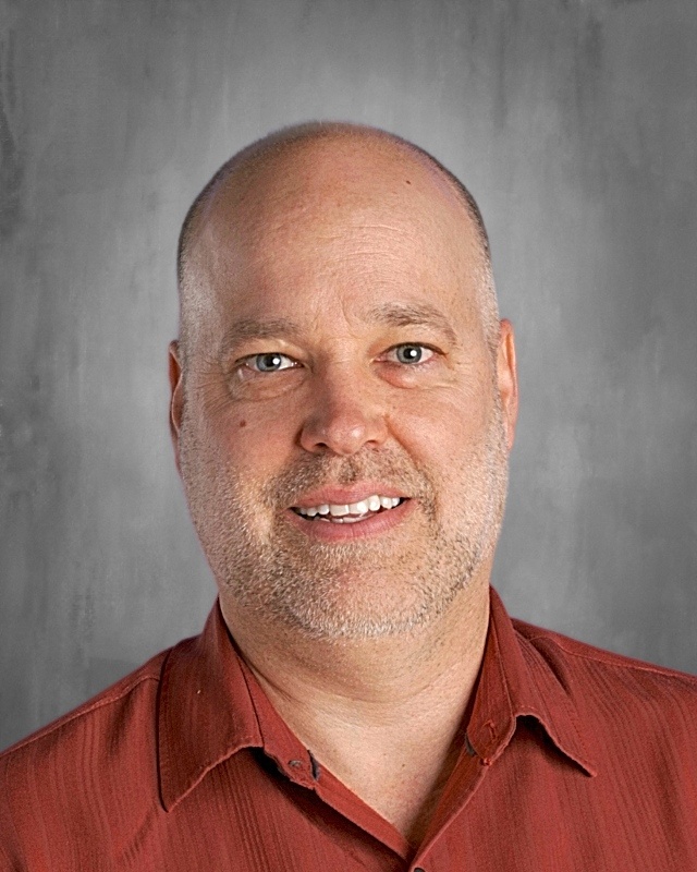 A person with a shaved head and beard, wearing a red shirt, smiles against a gray background.