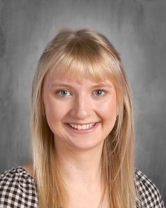 Smiling person with long blonde hair and bangs, wearing a checkered top against a gray background.
