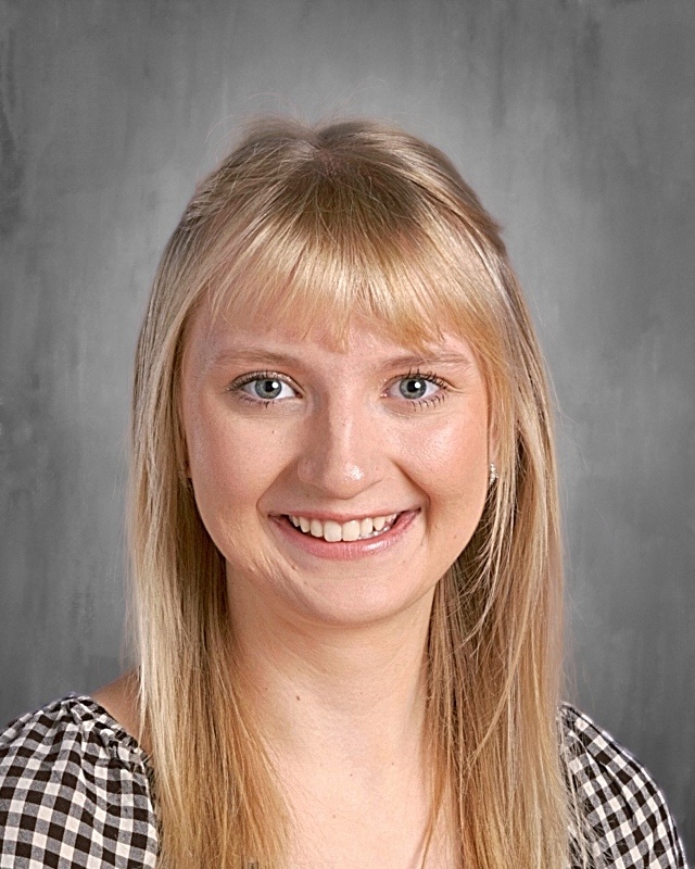 Smiling person with long blonde hair and bangs, wearing a checkered top against a gray background.