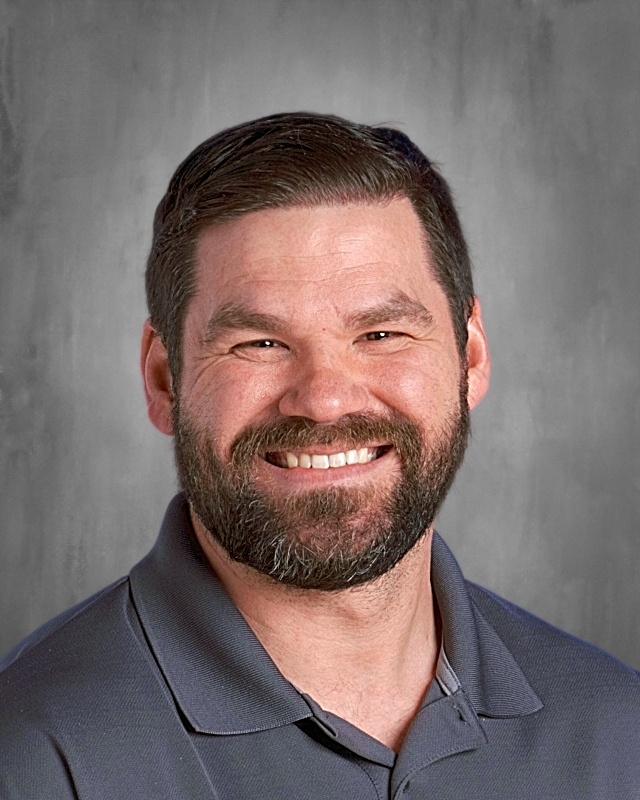 Smiling man with a beard wearing a gray polo shirt, set against a gray background.